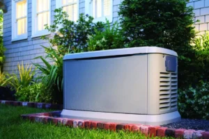 A standby Generator sits on a concrete pad beside a suburban house surrounded by green plants and lawn at dusk.