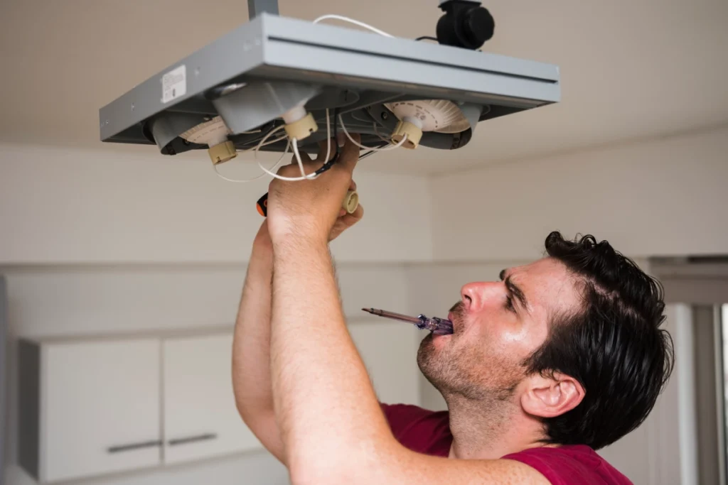 A man works on a ceiling light fixture with tools as part of end of year electrical maintenance.