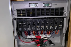 Open electrical panel showing a transfer switch with multiple circuit breakers, wiring, and two small meters at the top.